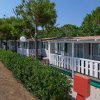 Filare di bungalow bianchi con verande, siepe verde lungo un vialetto sterrato.