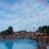 Persone intorno a una piscina guardano palloncini rosa e blu che volano in cielo.