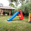 Area giochi con scivolo blu e altalene, circondata da alberi.