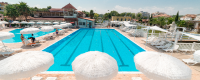 Piscina rettangolare con bordo in pietra, ombrelloni bianchi e edifici sullo sfondo.