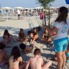Un gruppo di bambini ascolta una donna sulla spiaggia, circondati da ombrelloni e sedie a sdraio.