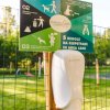 Cartello con regole per l'area cani e un dispenser su un palo in un parco verde.